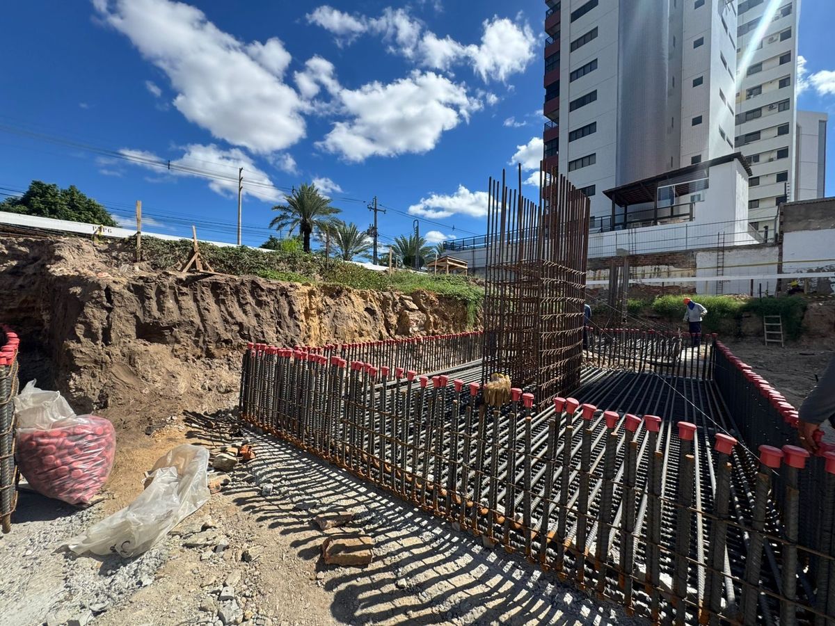 Armação de aço das fundações vista do chão, com as pontas dos vergalhões protegidas por capas vermelhas e edifícios ao fundo.