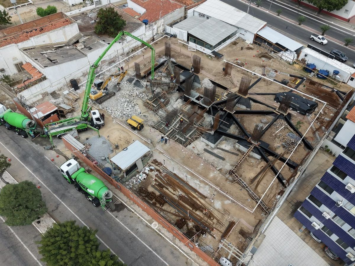 Vista de drone do canteiro com caminhão-bomba e betoneiras verdes junto às vigas baldrame.