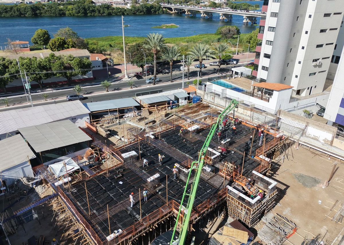 Vista de drone da concretagem da laje do pilotis, com operários e a lança verde do caminhão-bomba; ao fundo, o rio e a ponte.