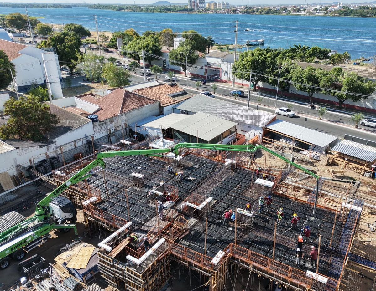 Vista de drone da laje do pilotis em concretagem, com a avenida da orla e o Rio São Francisco ao fundo.